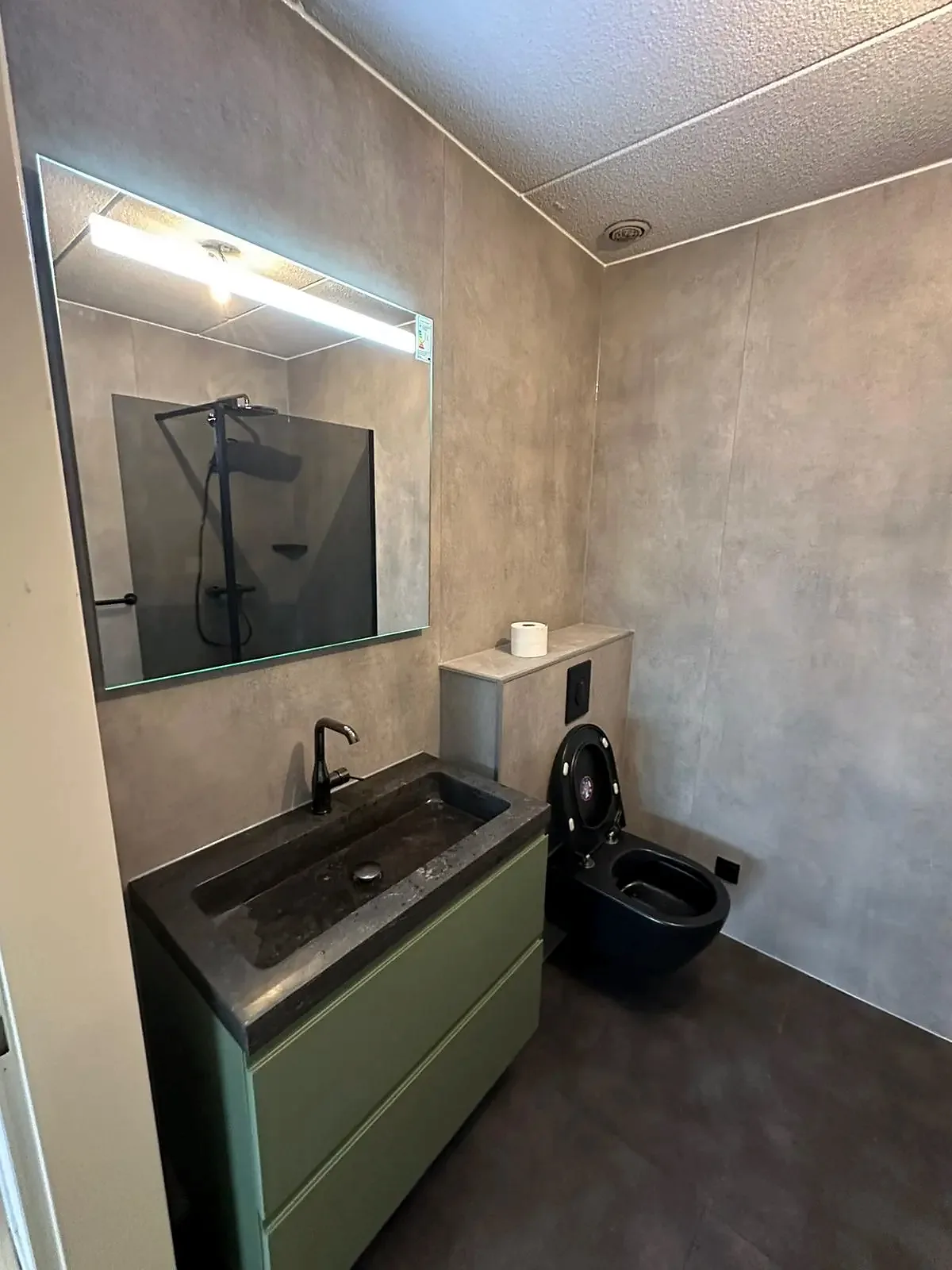 Betonlook wandpanelen met zwart hangtoilet, groen wastafelmeubel en stenen wastafel