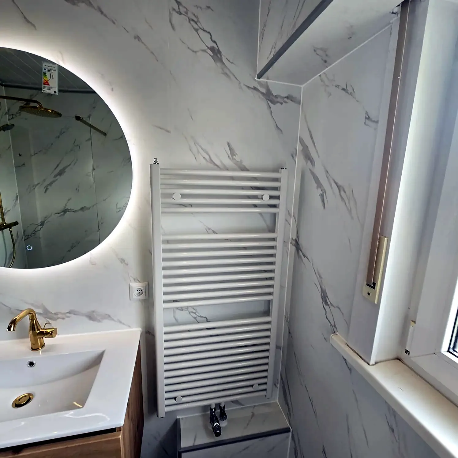 Witte marmerlook wandpanelen met ronde verlichte spiegel, eiken wastafelmeubel en gouden kraan